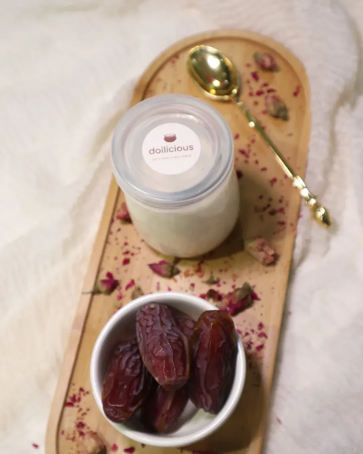 A jar of Doilicious mishti doi on a bamboo tray with a bowl of dates, rose petals and a gold spoon