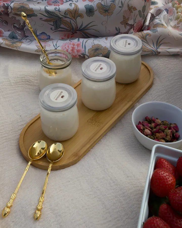 Four jars of mishti doi on a bamboo tray with gold spoons, rose buds and strawberries on a floral picnic blanket
