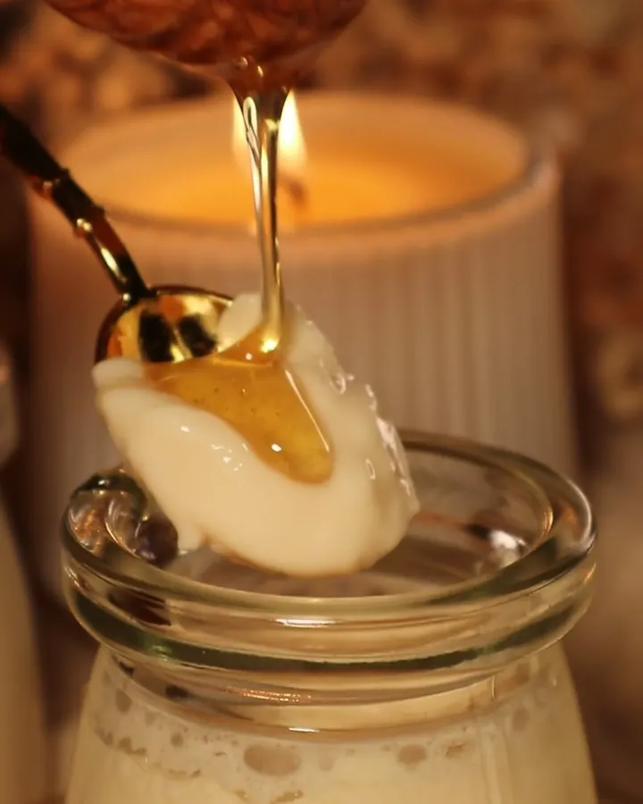 Honey pouring from a wooden dipper onto a spoonful of mishti doi above a jar, candlelight behind